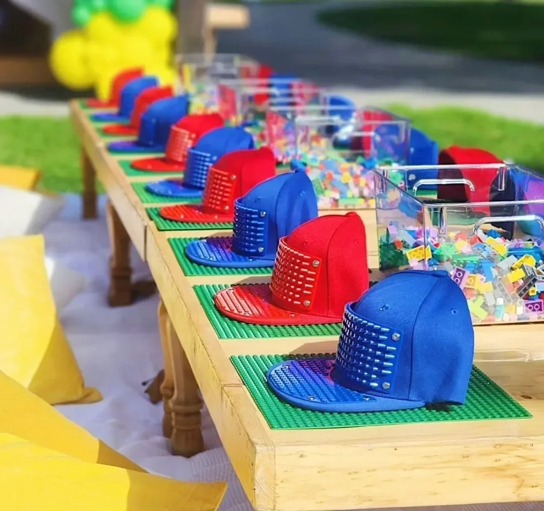 Table with red and blue baseball caps, colorful LEGO bricks, and plexiglass containers filled with small LEGO pieces. kids soiree craft activity parties