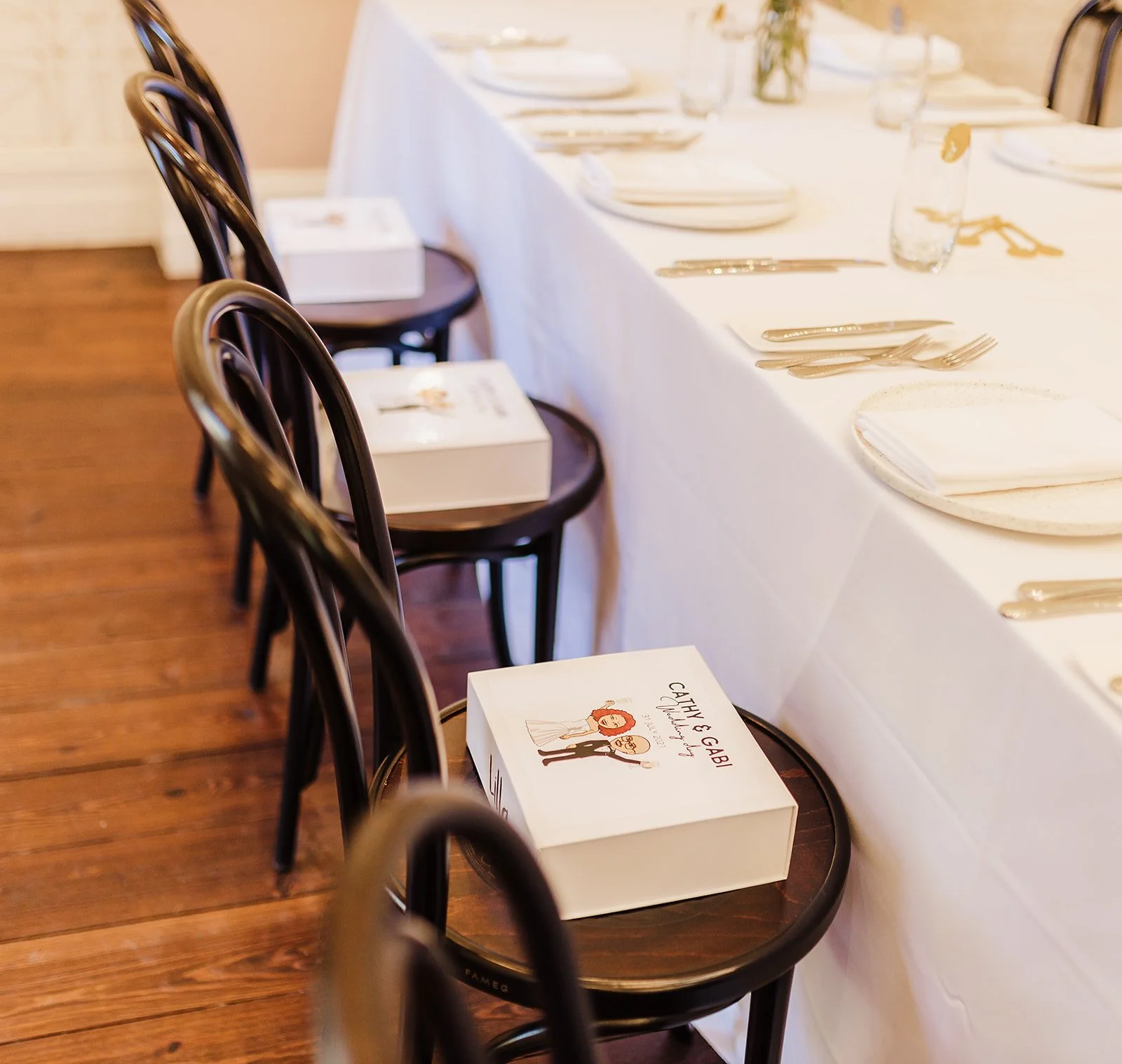 A long dining table set with white napkins, plates, forks, knives, and glasses, with black chairs around it, and a small cake box on one of the chairs. kids soiree craft activity parties