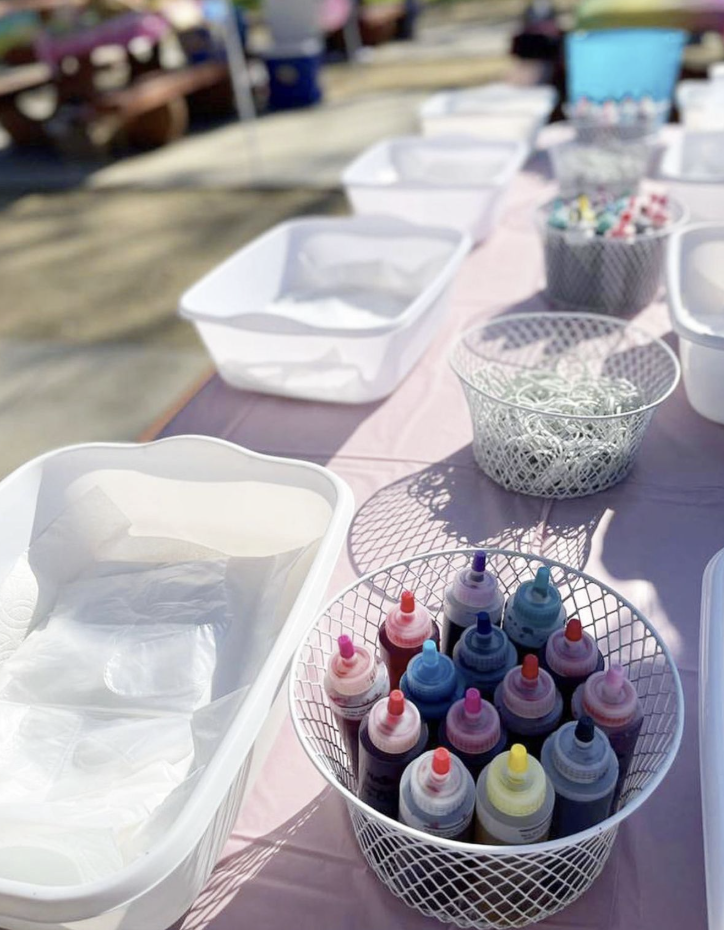 Colorful bottles of cake or cookie icing in a wire basket, outdoors table setup with empty containers and bowls, likely for a baking or decorating activity. kids soiree craft activity parties