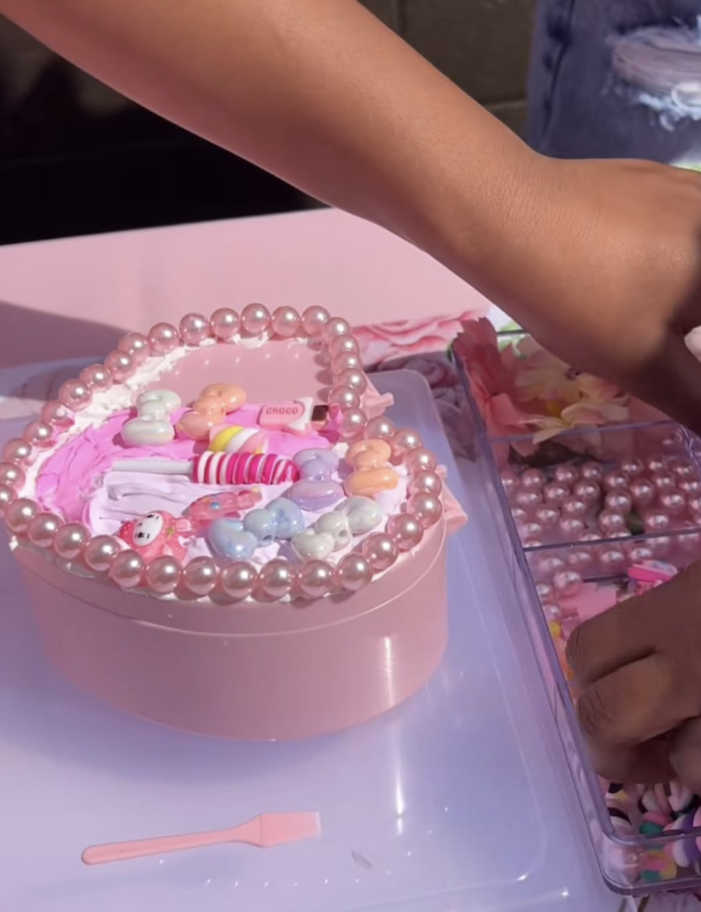 A pink cake decorated with pearl-like beads, candy, and sprinkles, placed on a table with hands reaching into a container of colorful candies nearby. kids soiree jewellery box decoden party