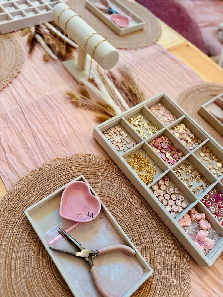 Jewelry-making workspace with a pink felt cloth, organized compartments of various beads, charms, and findings, a jewelry clamp, and pink ceramic dish. kids soiree craft activity parties