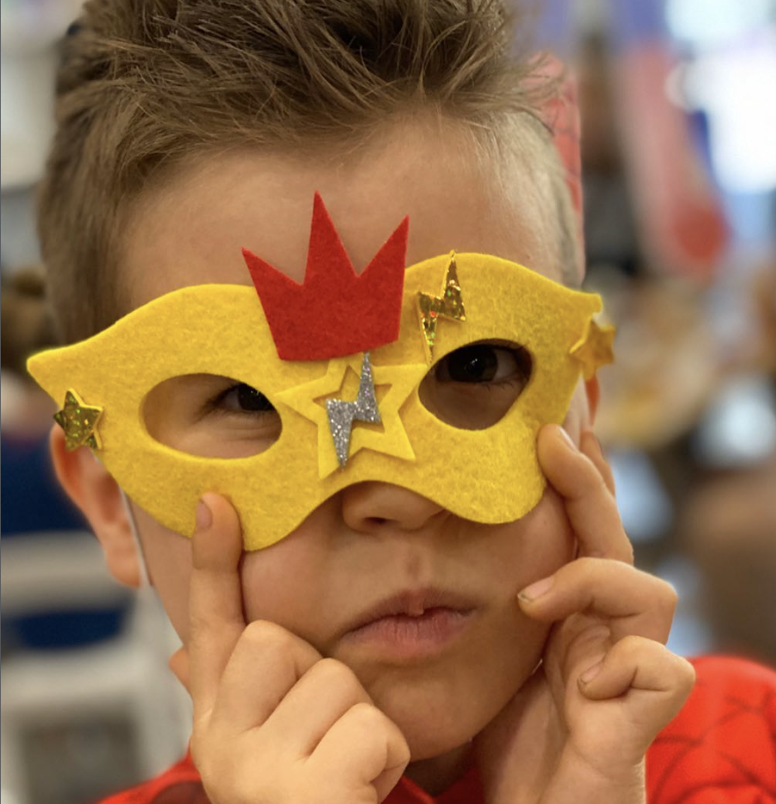 Child wearing a yellow felt superhero mask decorated with a red crown, stars, and lightning bolts, peering through the mask with curiosity. for a kids soiree party