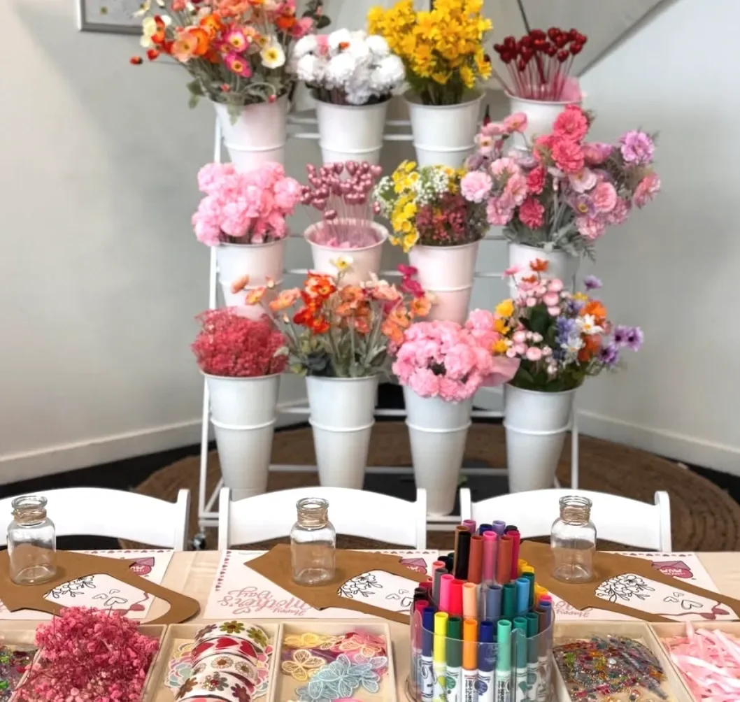 A display of colorful flowers in white pots arranged on a tiered stand, with a table in the foreground holding empty jars, colorful markers, and decorated paper items for a celebration or craft activity. kids soiree craft activity parties