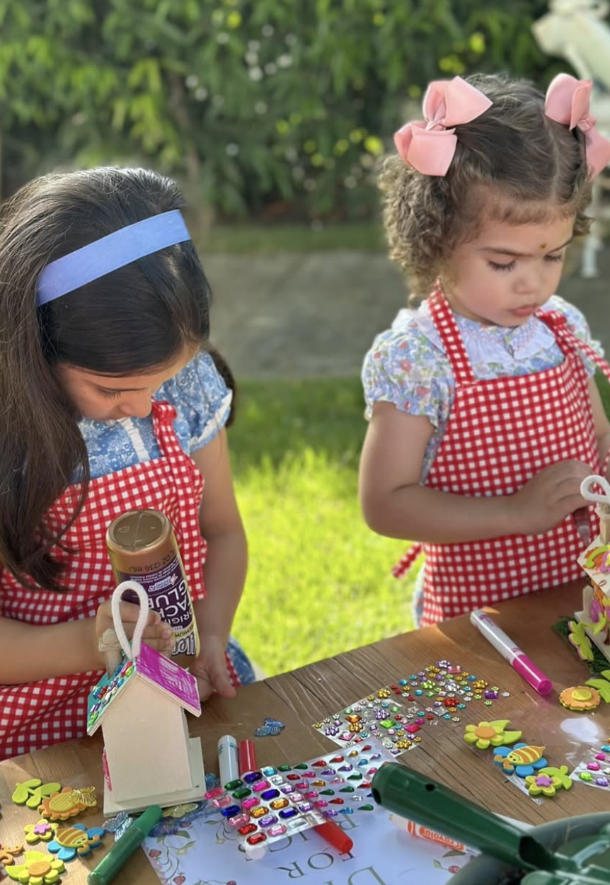 Two young girls with curly and straight hair, wearing red and white checkered aprons, engaging in arts and crafts outdoors. kids soiree craft activity parties