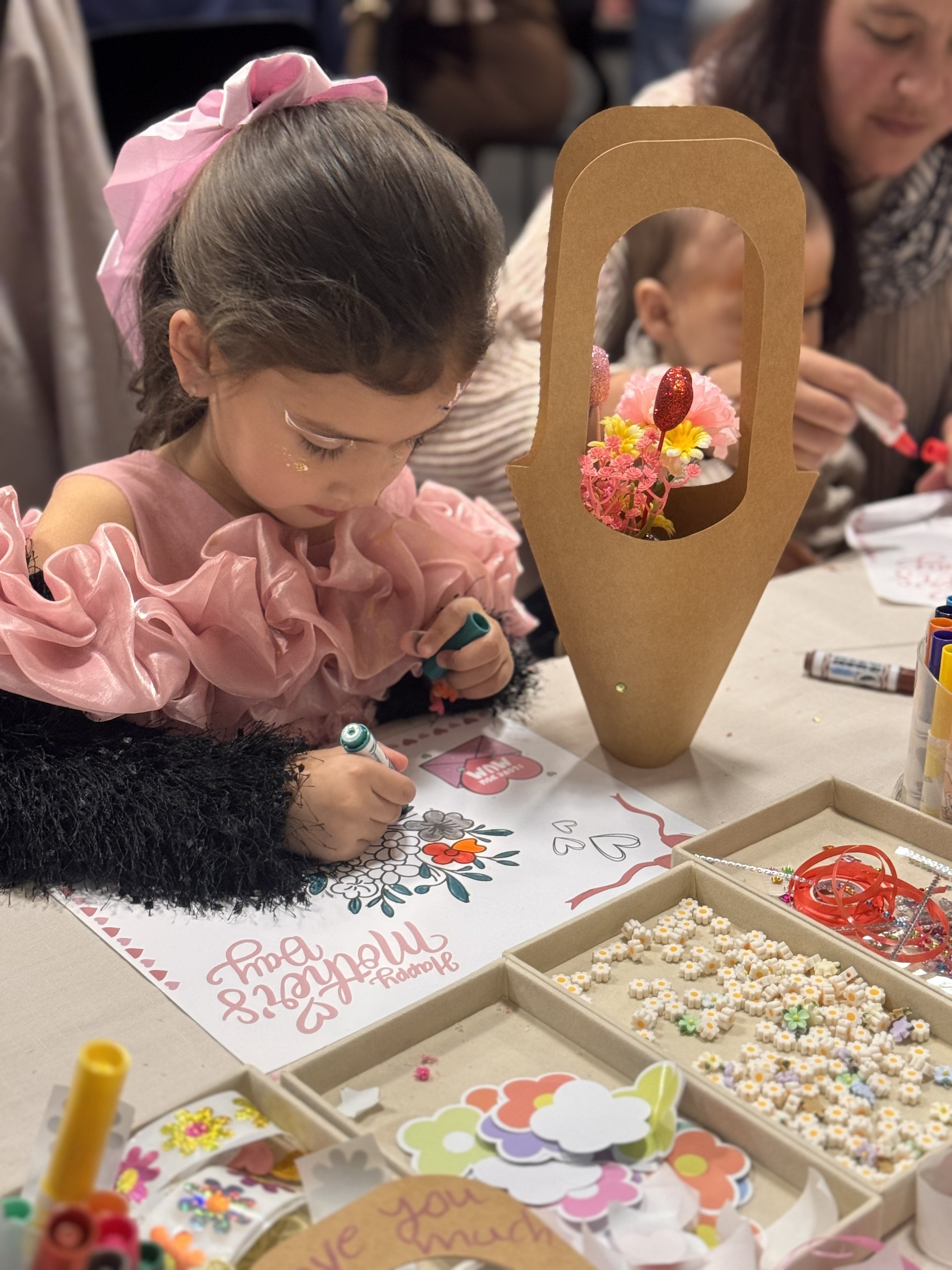 A young girl with a pink bow in her hair and wearing a pink dress is coloring a love letter with a marker. There are various craft supplies and decorative items on the table, kids soiree craft activity parties