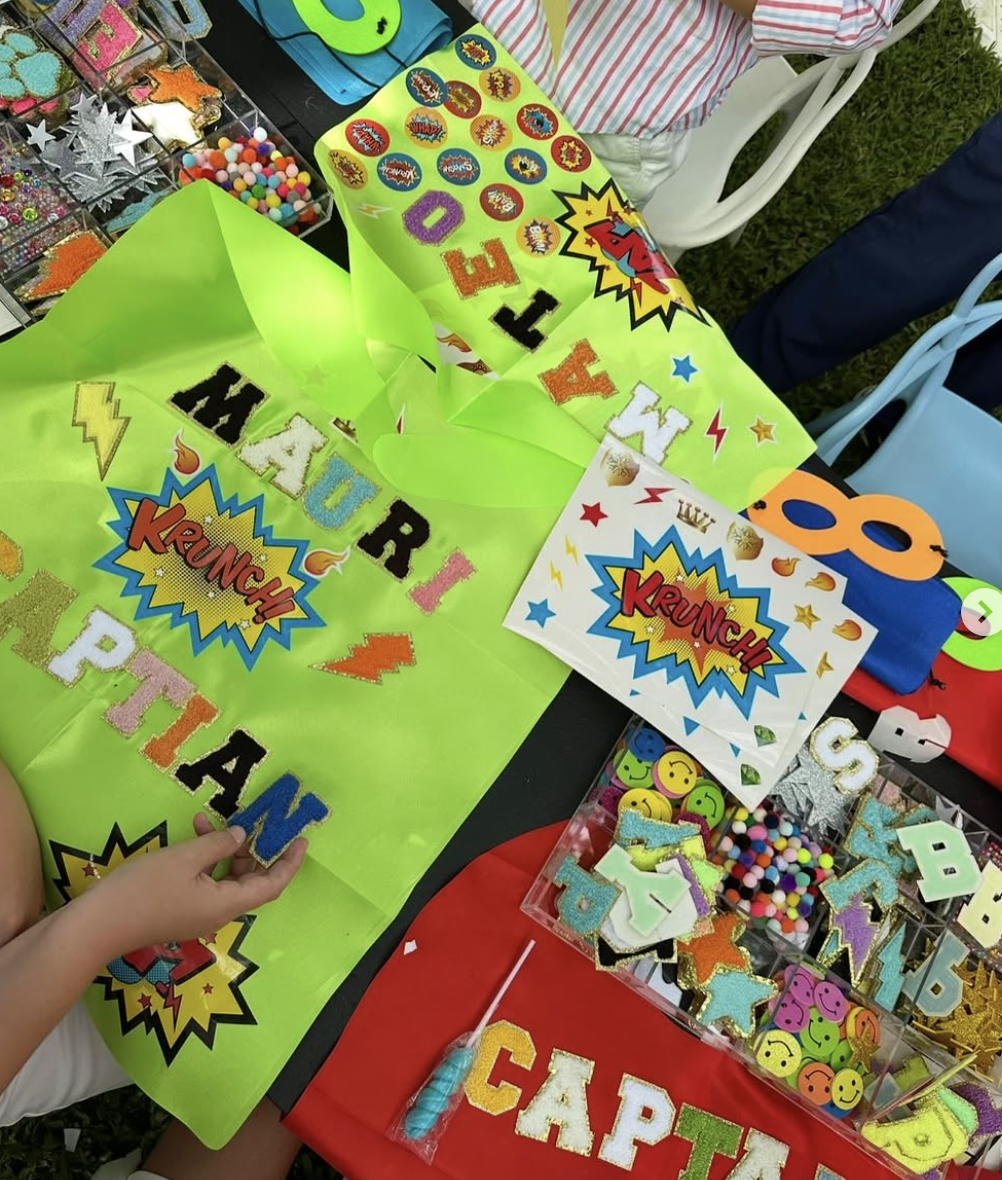 Colorful craft supplies and decorations on a table, including glitter letters, foam letters, stickers, pom-poms, and superheroes-themed items, likely for a birthday celebration. kids soiree craft activity parties