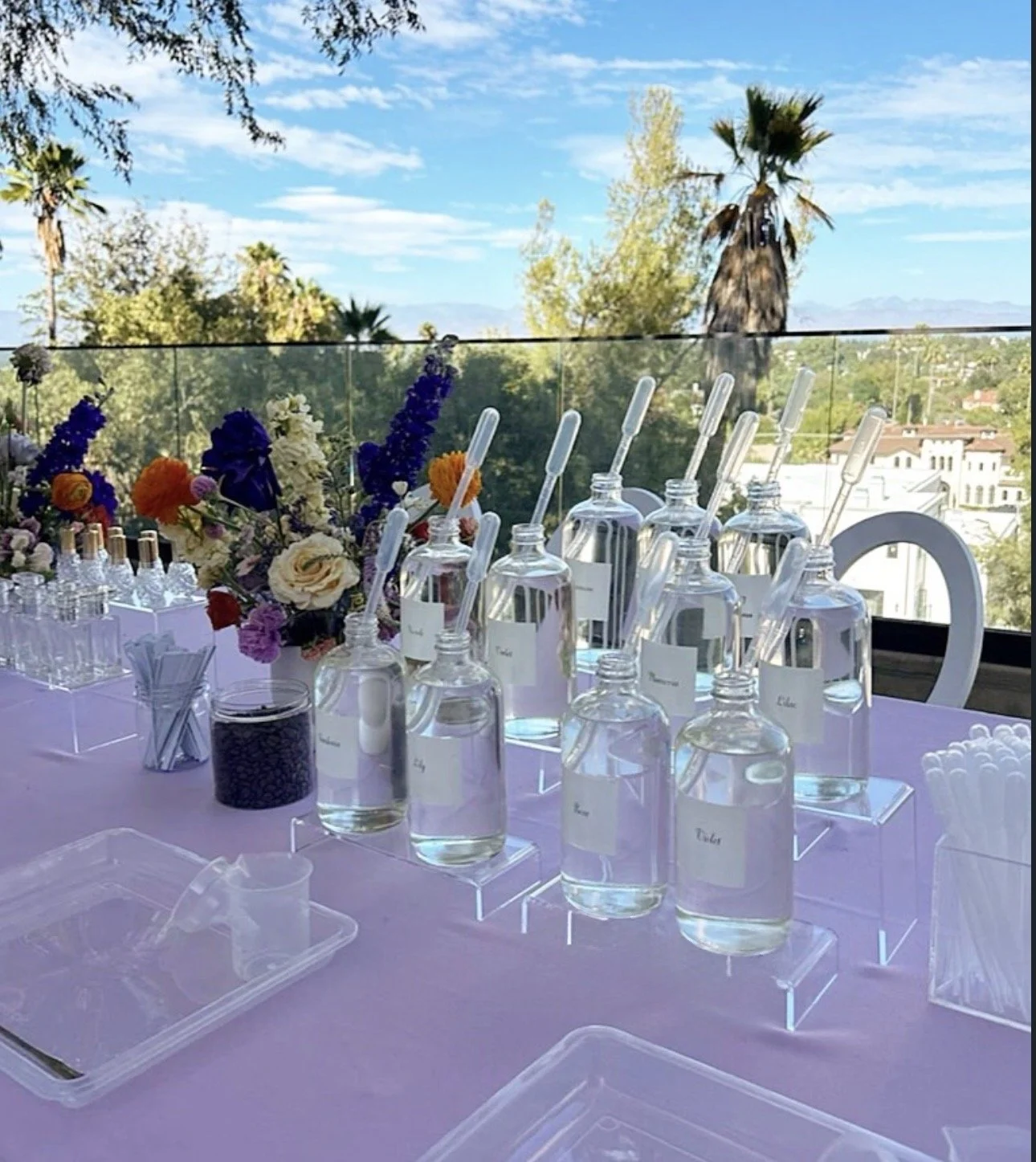 Decorative table setup with glass bottles and droppers, a bouquet of colorful flowers, and small containers, against an outdoor backdrop of trees and a blue sky. kids soiree craft activity parties