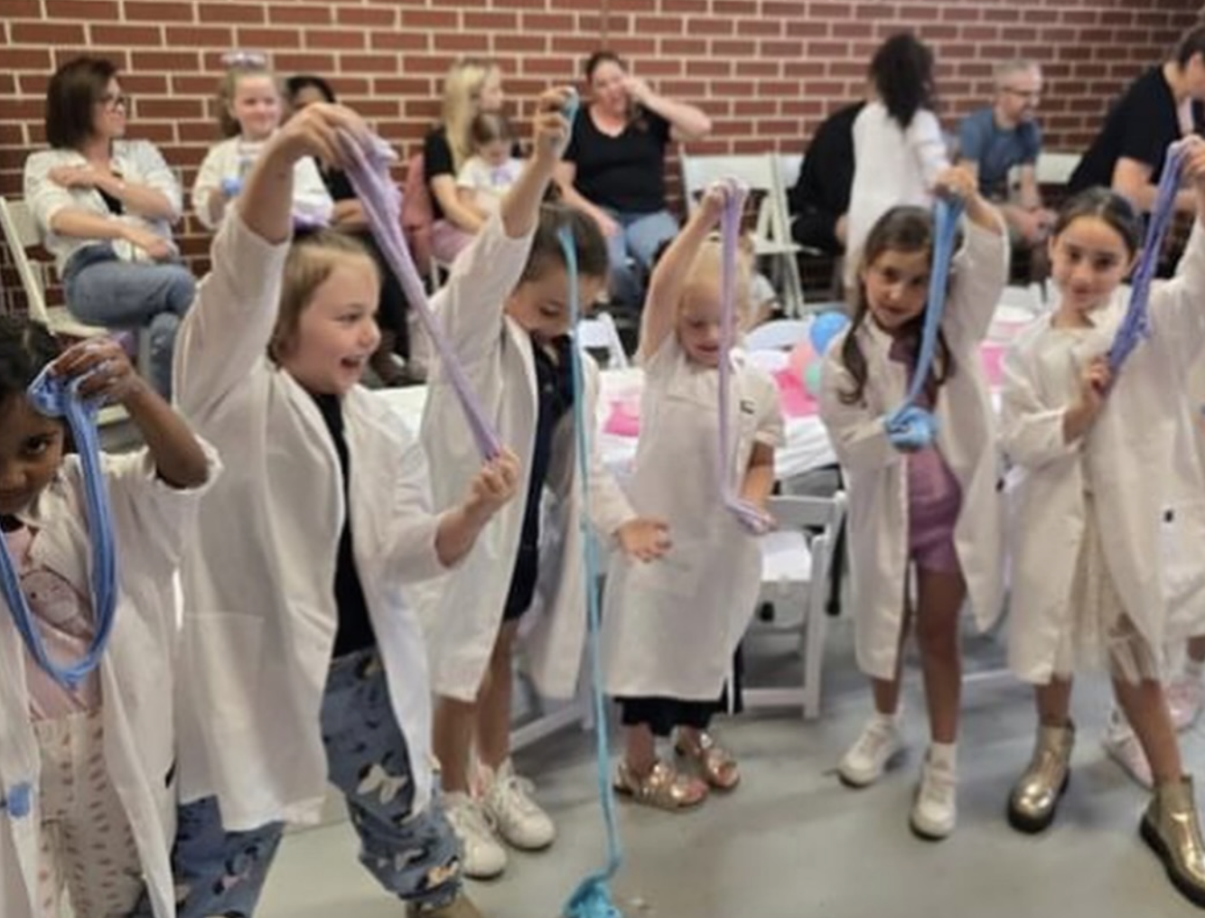 Children in white lab coats holding colorful slime, standing in a line in a science classroom. Kids Soiree Slime Party