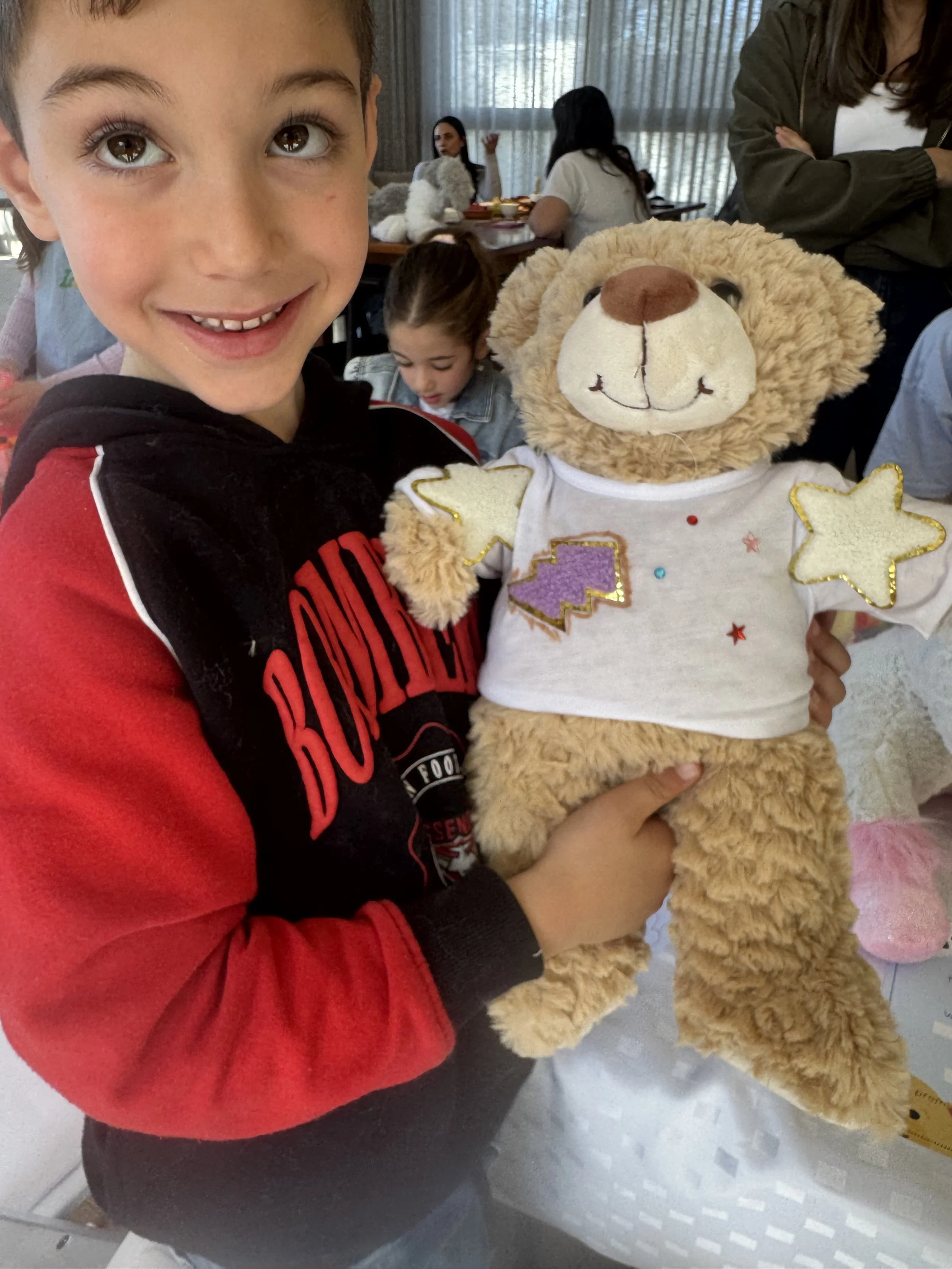 A young boy with brown hair and big brown eyes smiling while holding a stuffed lion toy. kids soiree adopt a teddy