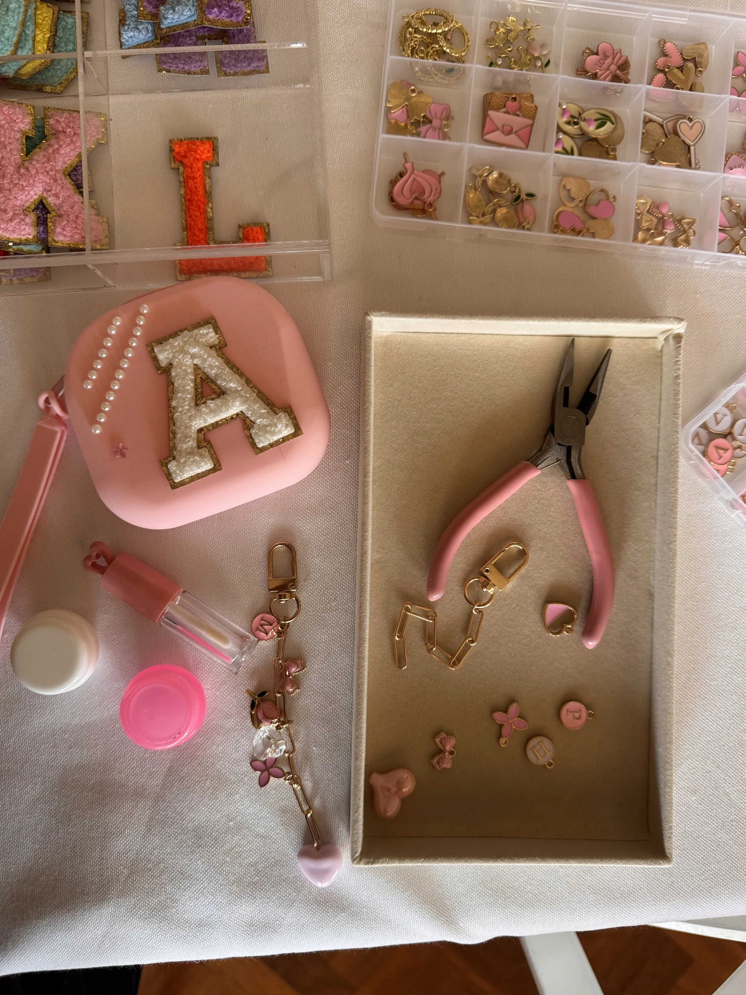 A collection of jewelry and accessories including a pink heart-shaped item, a pearl-edged letter 'A', gold and pink charms, and various decorative items, organized in small containers on a table. lip gloss making kids soiree