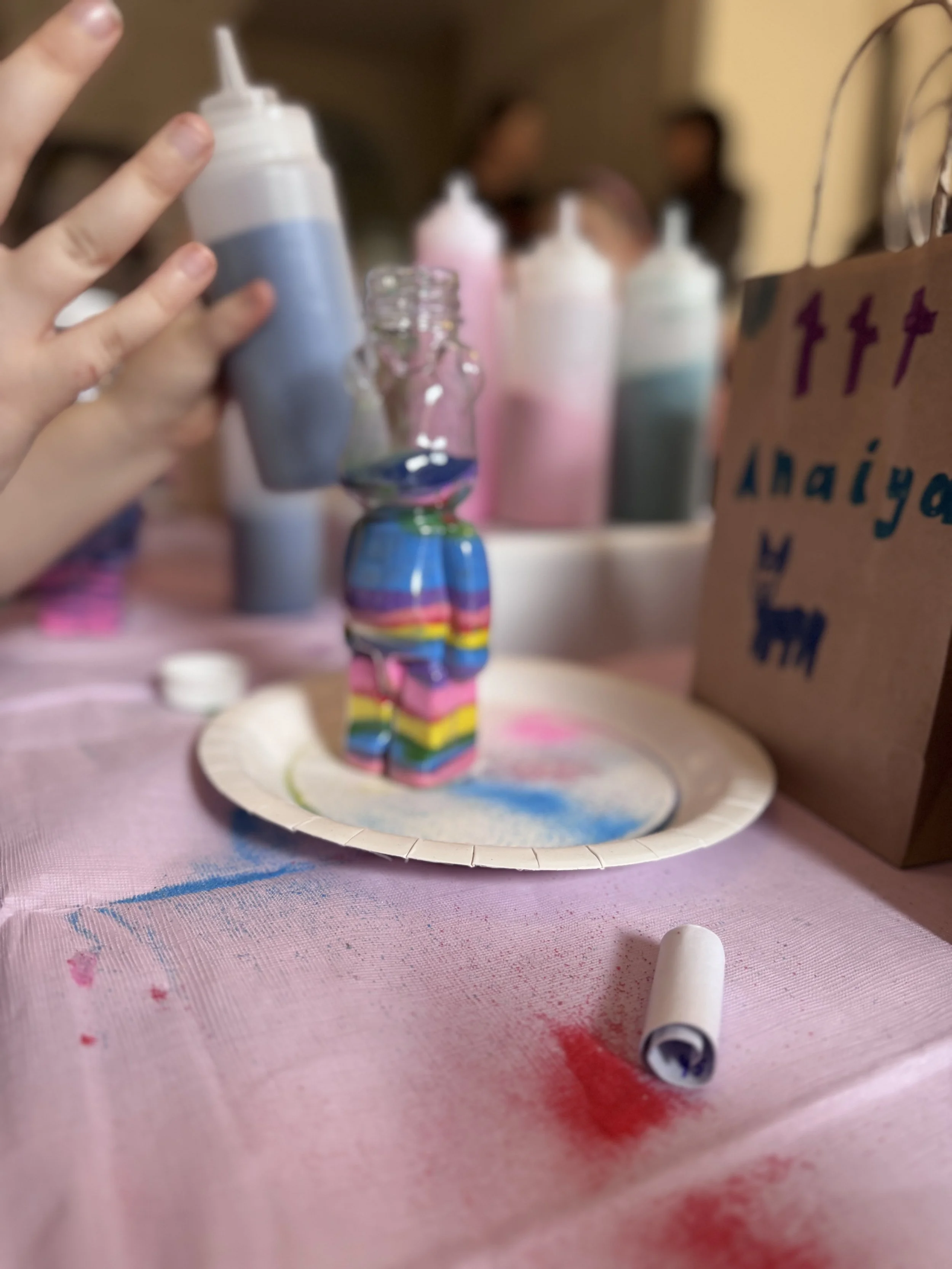 Close-up of a handmade rainbow-colored castle made from stacked cardboard boxes, with a pink and blue color scheme, on a pink craft table surrounded by art supplies and colorful bottles. kids soiree craft activity parties