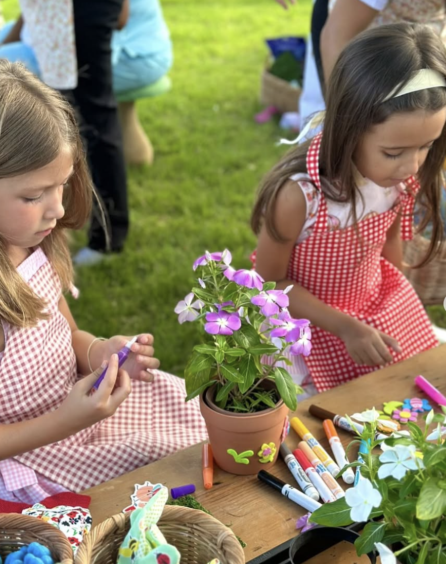 Two young girls in red and white checkered aprons sitting at a table outdoors, decorating flower pots with colorful markers, surrounded by plants, flowers, and craft supplies. kids soiree craft activity parties
