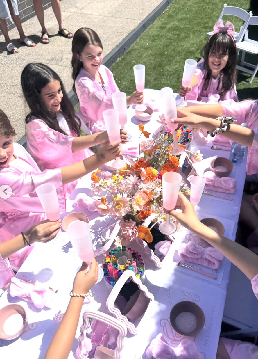 Children in pink dresses celebrating at a table with pink cups, colorful pens, and a flower arrangement. Kids Soiree Pamper Party