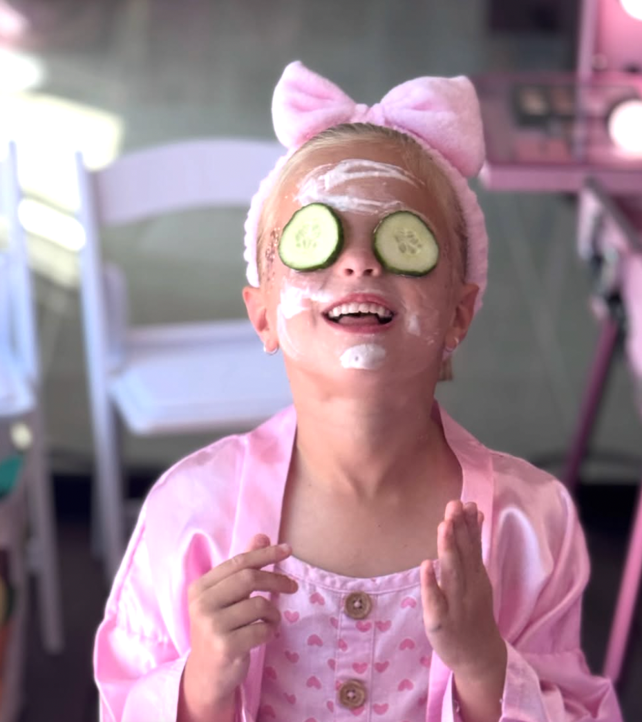 A young girl in pink pajamas with a large pink bow headband, smiling with hands raised, wearing cucumber slices on her eyes and with a white cream on her face. kids soiree craft activity parties