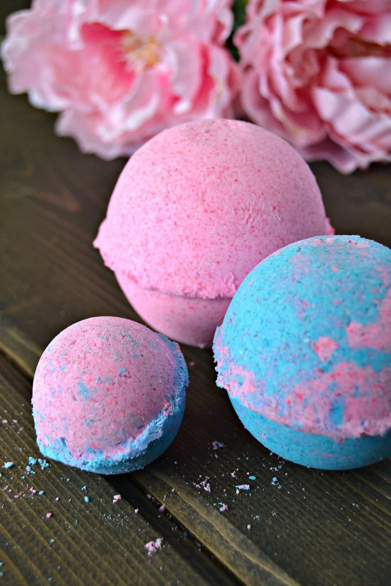 Close-up of colorful bath bombs, pink, blue, and purple, on a wooden surface with scattered powder and crumbs, with pink flowers in the background. kids soiree craft activity parties