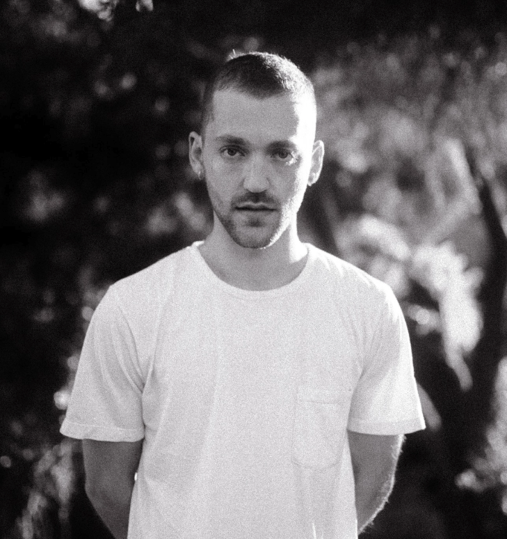 A young man with short hair and light facial stubble wearing a plain white t-shirt, standing outdoors with blurry trees in the background.