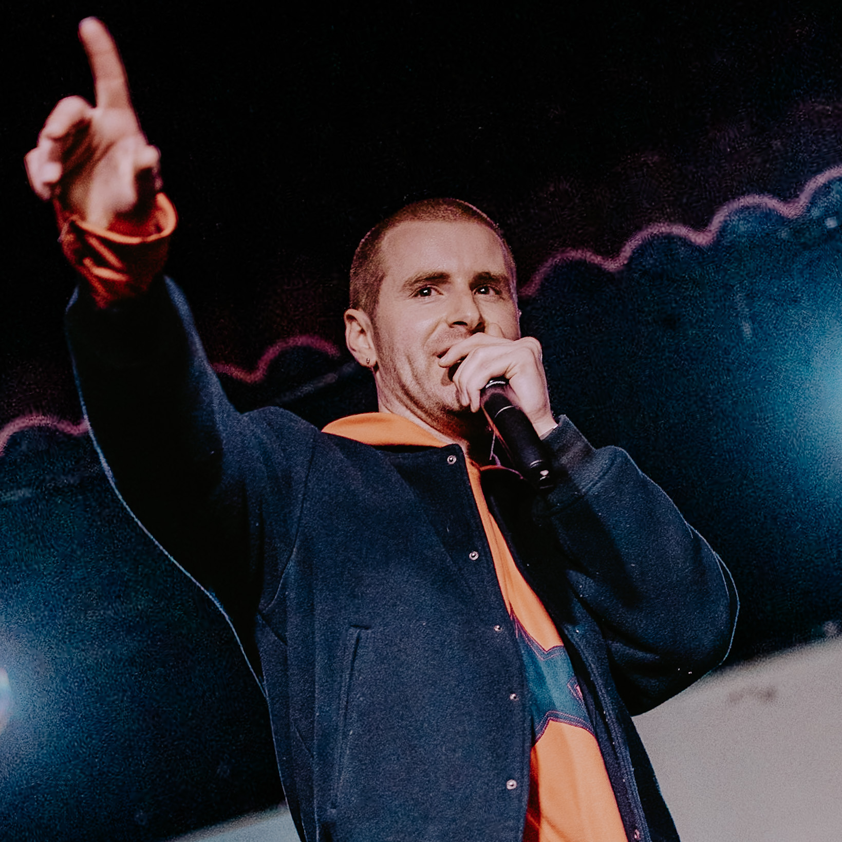 Young man with short hair speaking into a microphone, wearing a dark jacket over an orange shirt, with a pink zigzag line graphic in the background.