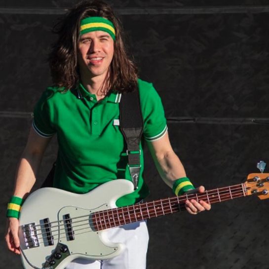 Person with long brown hair wearing a green polo shirt, green headband, and green wristbands, holding a white electric bass guitar.