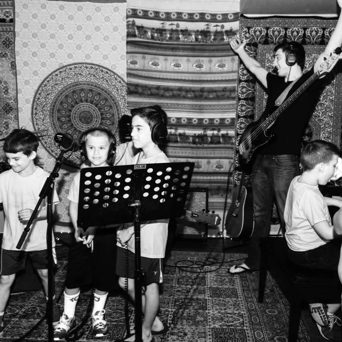 Group of children and a musician in a music studio; children wearing headphones, one girl with a microphone stand, and a boy playing a guitar with music notes on a music stand.