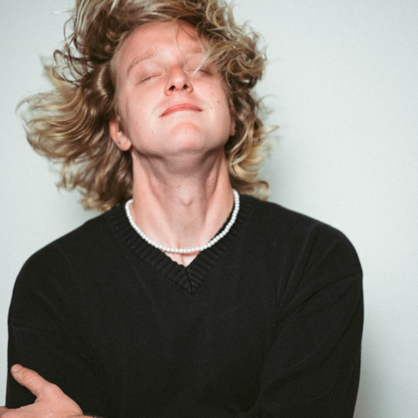 A woman with curly blonde hair, wearing a black sweater and pearl necklace, smiling with her eyes closed against a light background.
