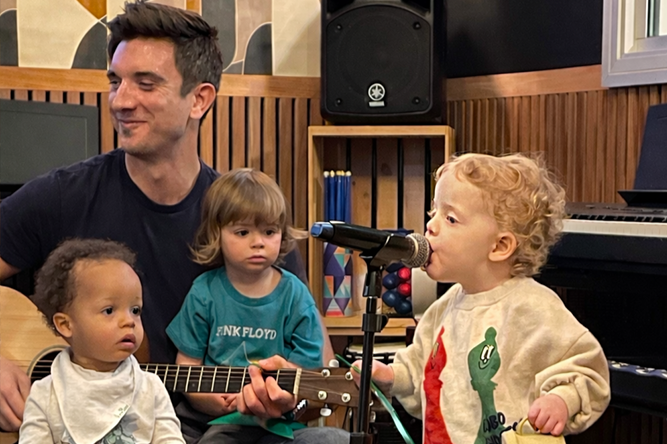 A young boy with curly blonde hair sings into a microphone, while an adult man and two young children observe. The man holds a guitar, and the scene appears to be in a music or therapy room with wooden walls, a speaker, and musical instruments.