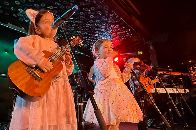 Two young girls singing and playing guitars on a stage with microphones, accompanied by two other performers, in a lively indoor venue.