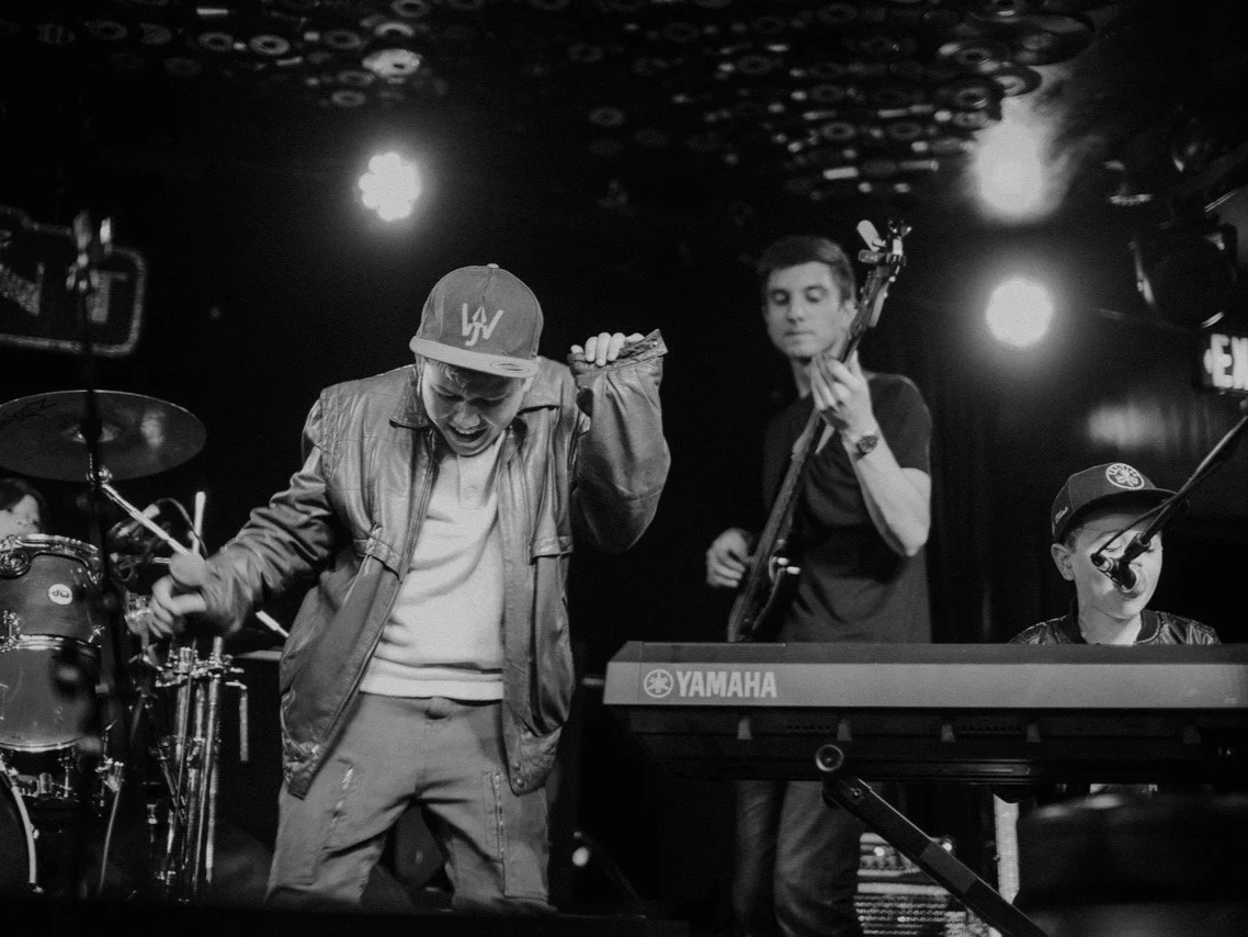 Musicians performing on stage, featuring a drummer, a singer, a bassist, and a keyboard player, with stage lights overhead.