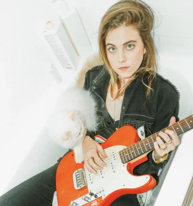 A woman with long, wavy hair holding an orange electric guitar, sitting in a white room with a fluffy white cat resting on her lap, looking directly at the camera.