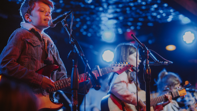 Three children performing on stage with guitars and microphones, illuminated by stage lights.