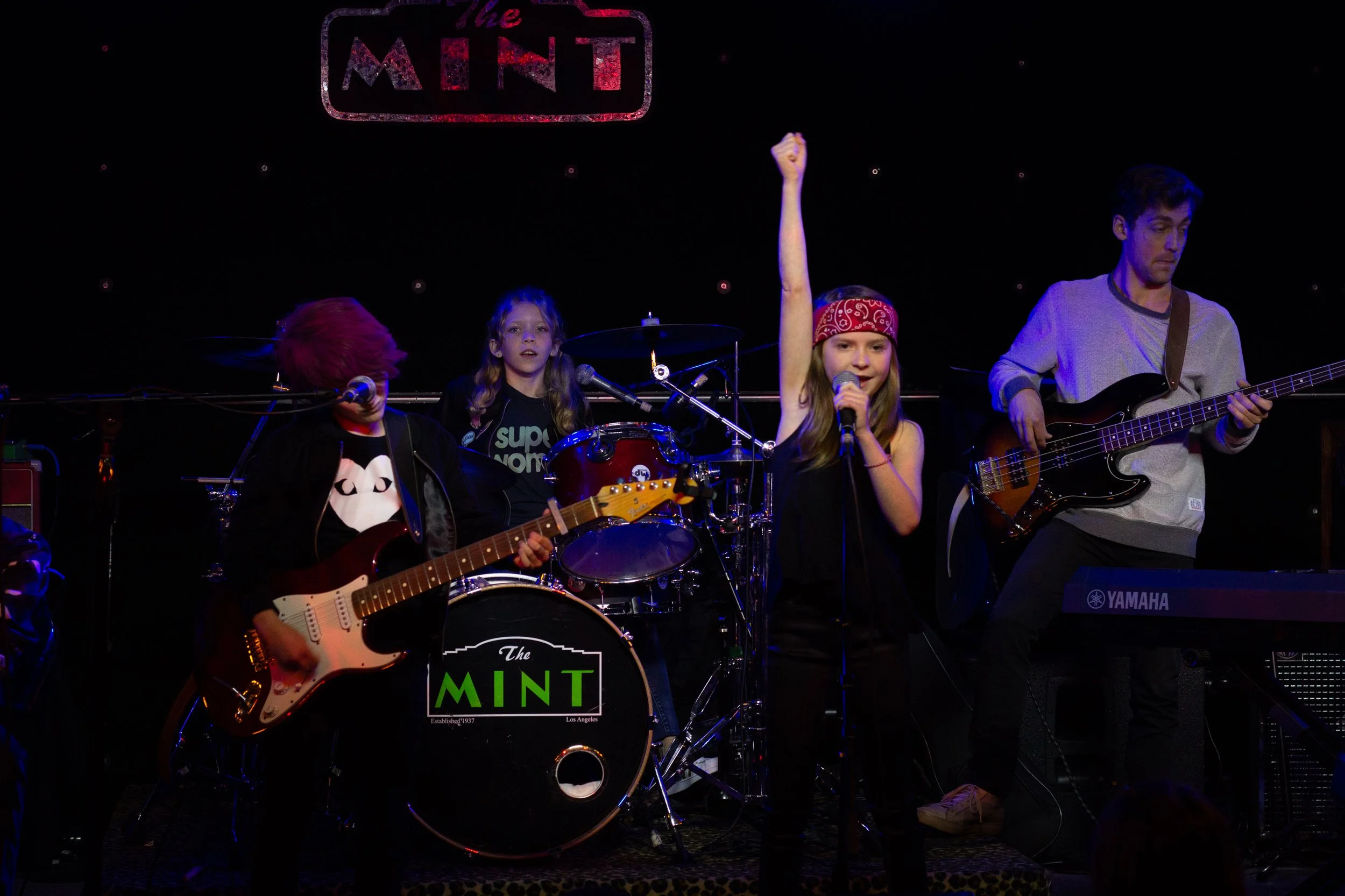 A band performing on stage with three young musicians playing guitars and drums, and a singer with her arm raised, at The Mint music venue.