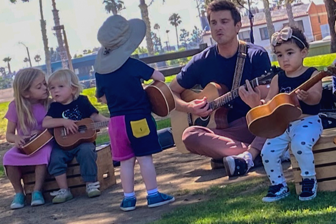 A man and four young children sitting outdoors, playing and teaching guitar.