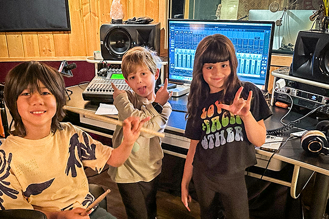 Three kids in a recording studio with sound equipment and a computer screen displaying audio mixer software, all making peace signs.