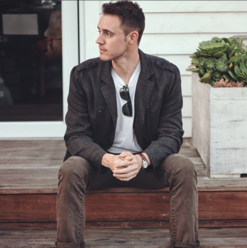 A young man in a dark gray jacket, white shirt, and brown pants sitting on a wooden porch, with sunglasses hanging on his shirt and a watch on his left wrist, looking to the left. There is a large white planter with succulents on his right side and a glass door behind him.