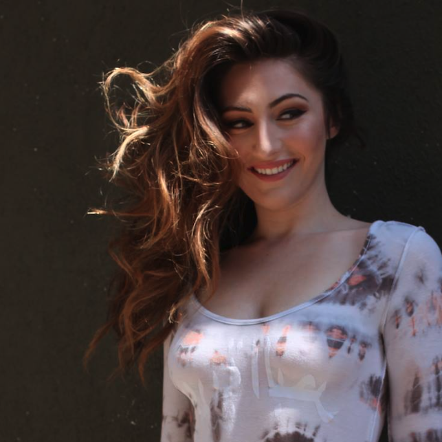 A woman with long, wavy brown hair and a light complexion, smiling and looking away from the camera, wearing a fitted white top with a colorful abstract pattern, standing outdoors against a dark background.
