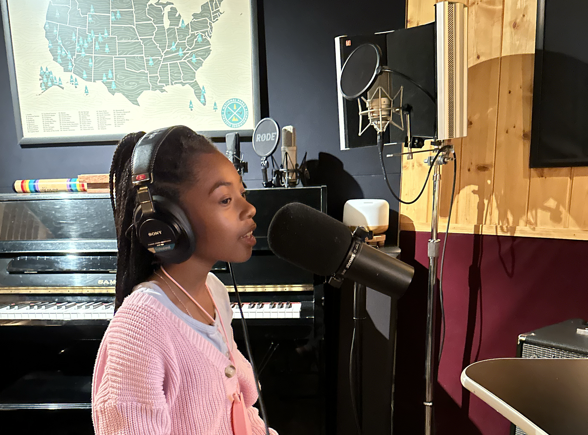 A young girl with braided hair wearing headphones, singing into a professional microphone in a music studio. Behind her, there is a black Steinway piano, a large map of the United States on the wall, and recording equipment.