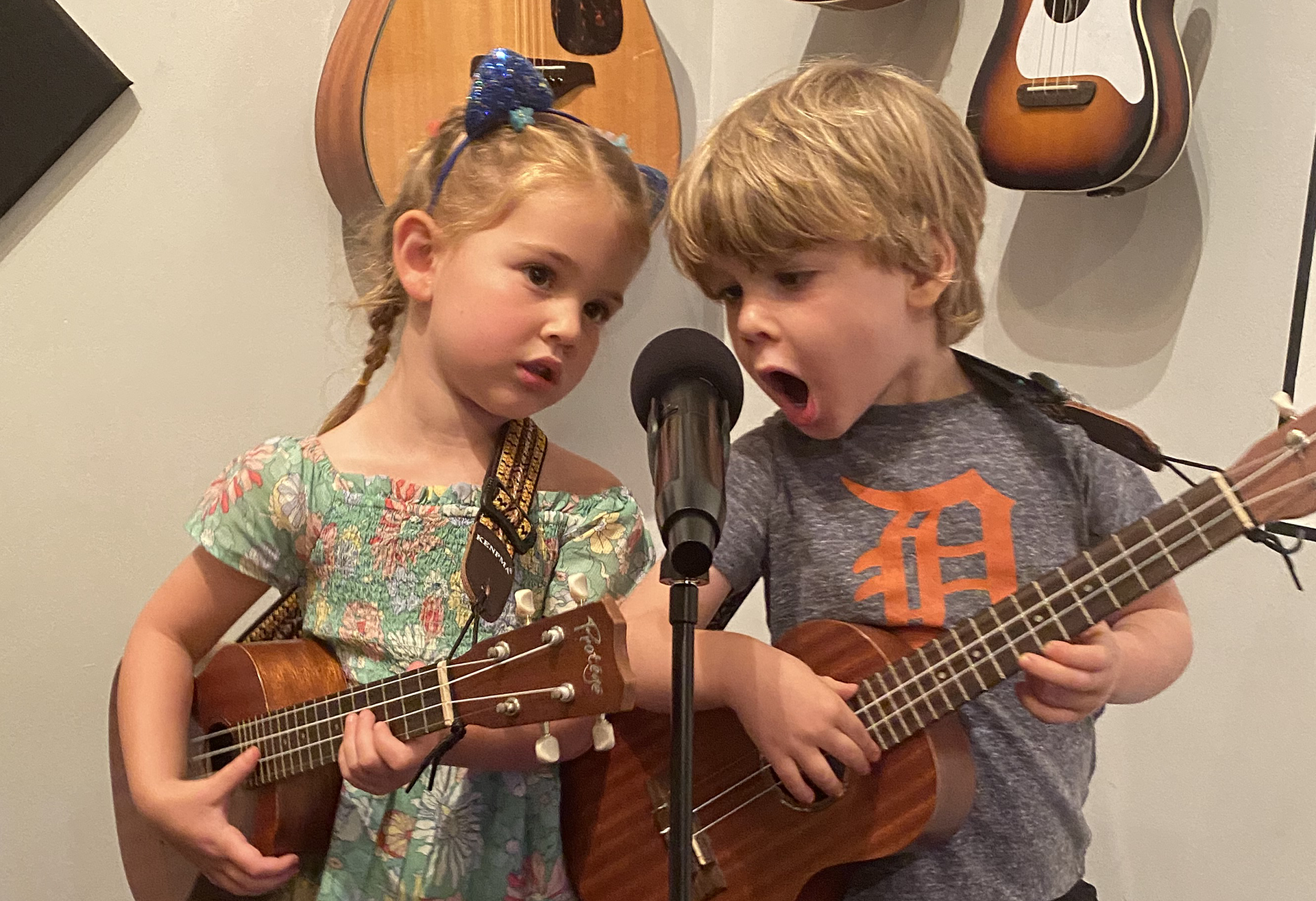 Two young children, a girl and a boy, are playing guitars and singing into a microphone, standing close together in front of a wall with two guitars hanging on it.