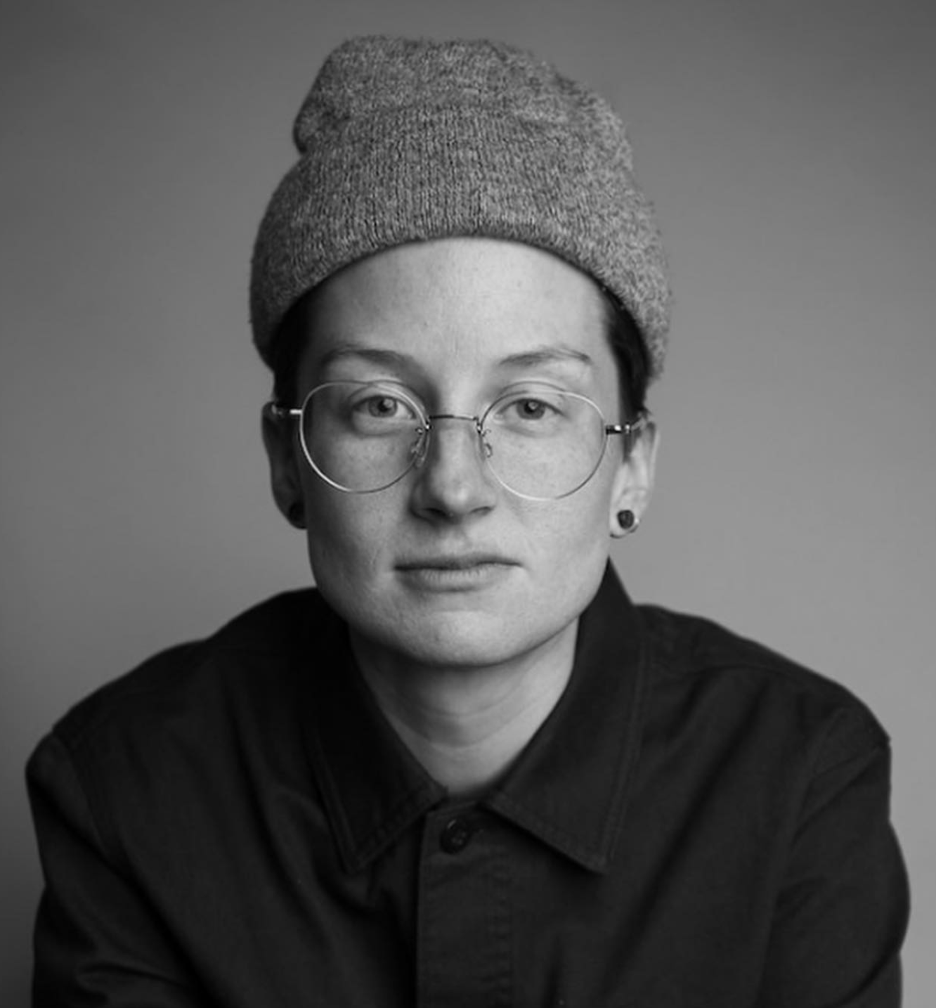 Black-and-white portrait of a woman with short hair, glasses, earrings, wearing a beanie and a dark buttoned shirt against a plain background.