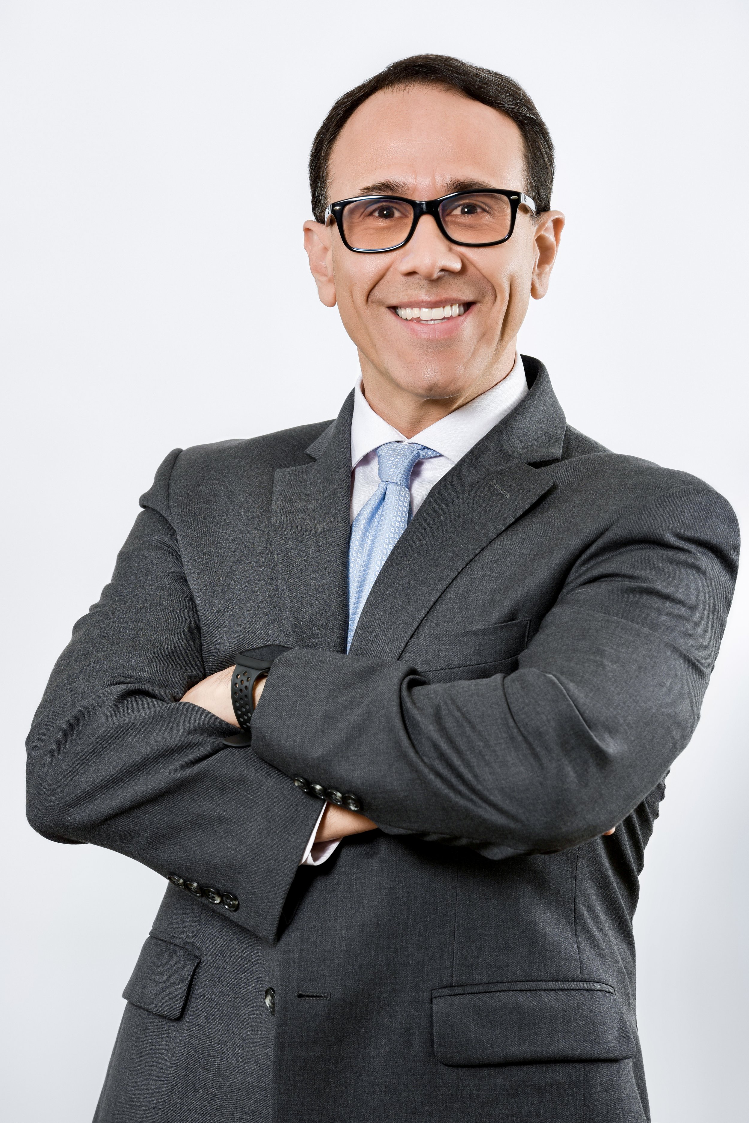 A man in a dark gray suit, white shirt, and light blue tie, smiling with glasses, arms crossed, against a plain white background.