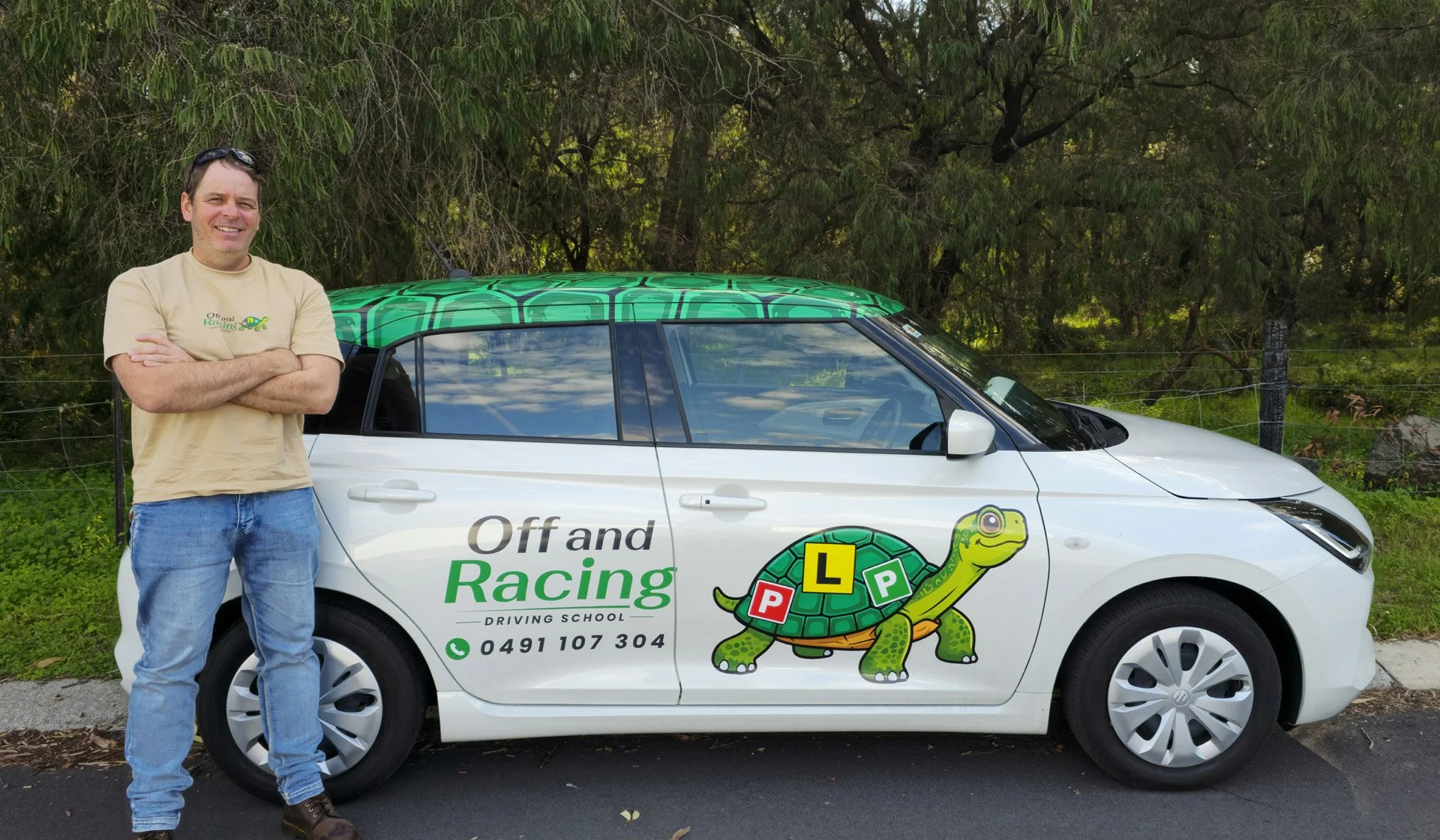 Off and Racing Driving School Bunbury.  A man standing with arms crossed in front of a white driving school car that has green and yellow dinosaur graphics and driving school information. The man is smiling, wearing a beige T-shirt and blue jeans.