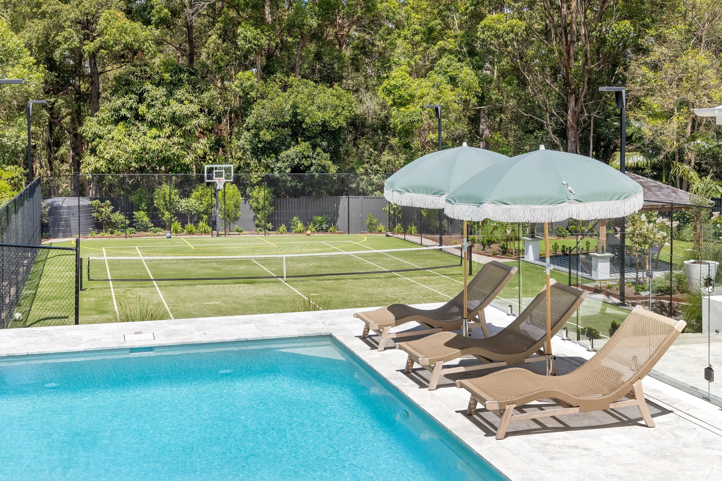 A backyard swimming pool with lounge chairs, shaded by a large patio umbrella, overlooking a grassy sports court surrounded by trees.