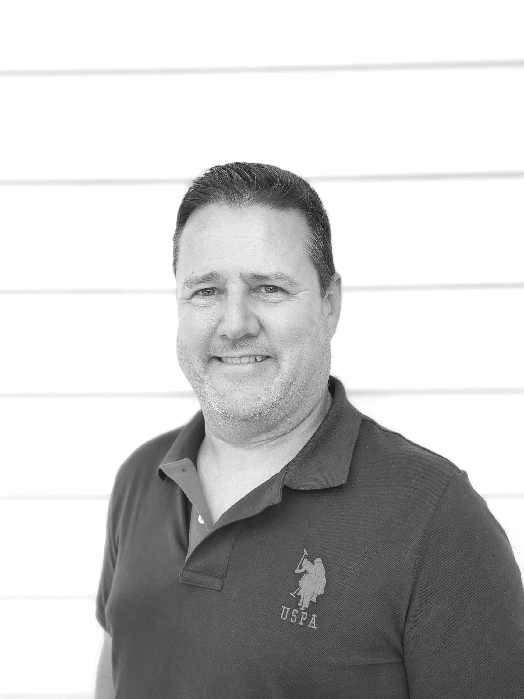 Black and white portrait of a middle-aged man with dark hair, smiling, wearing a collared shirt with a Polo Ralph Lauren logo, standing against a white background with horizontal lines.