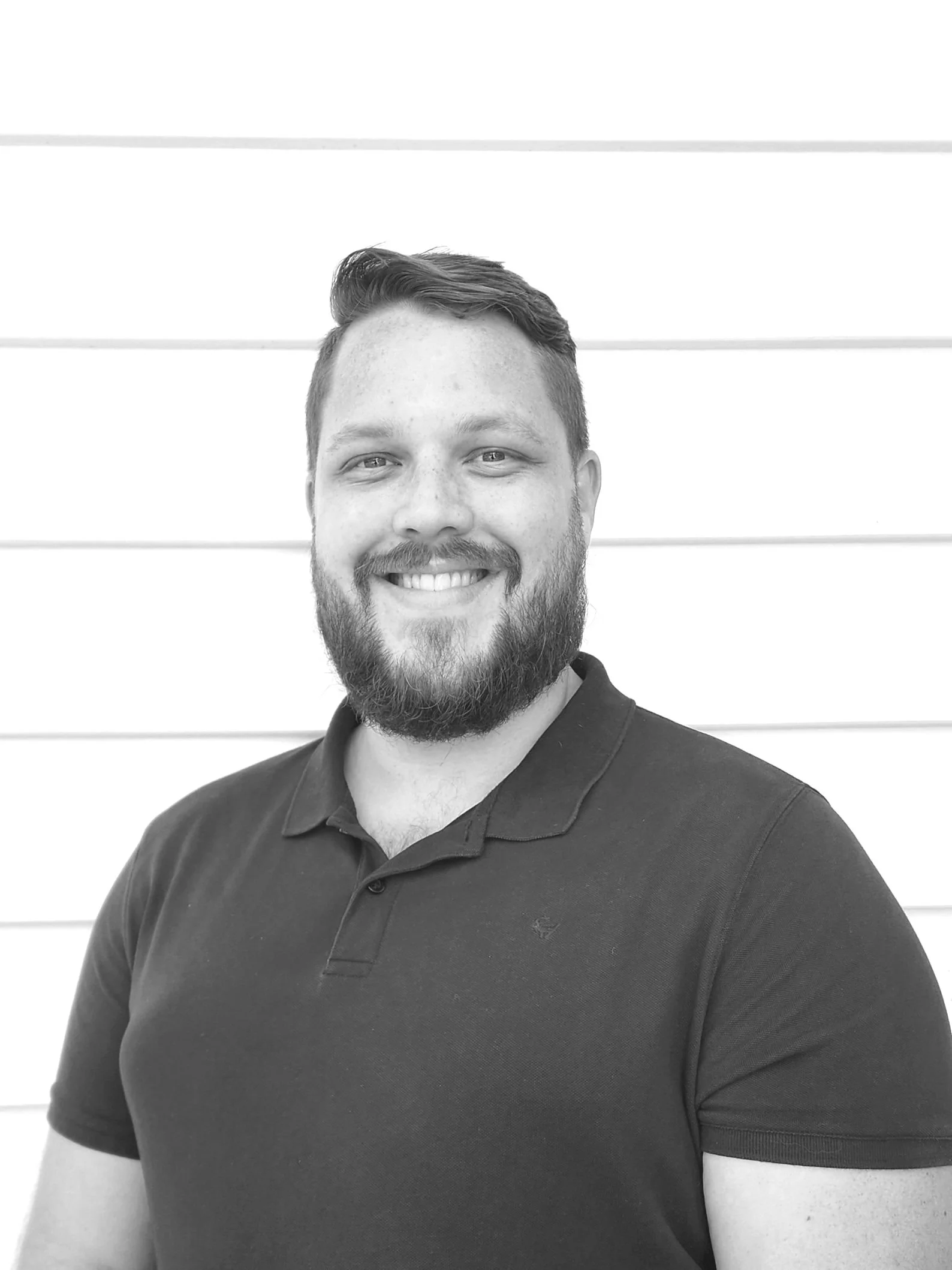 Black and white portrait of a man with a beard smiling, wearing a polo shirt, against a horizontally paneled background.