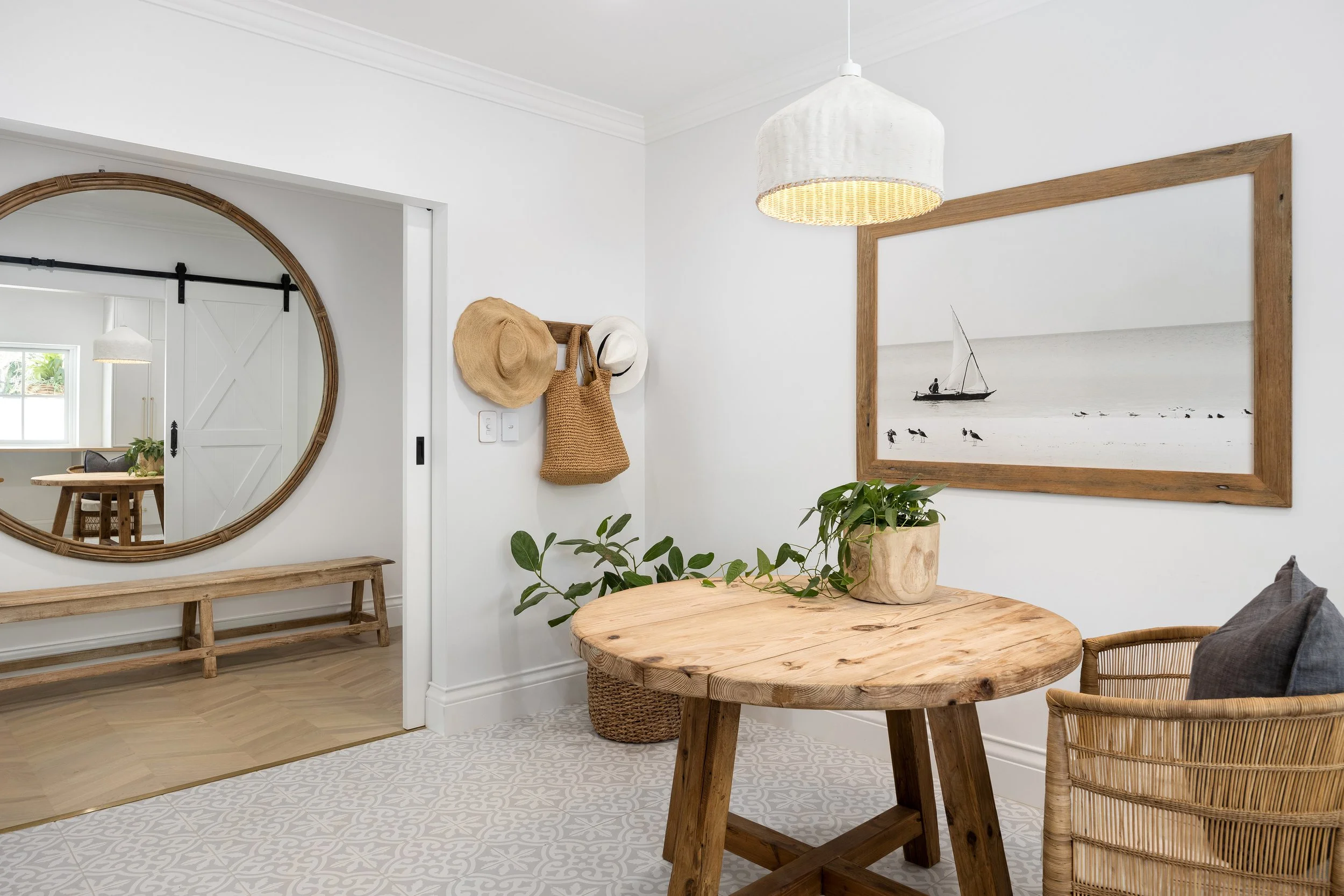 Interior view of a home with a round wooden table, a wicker chair, a basket with a plant, and a large framed black-and-white nautical painting on the wall. A mirror with a rattan frame reflects a doorway with a sliding barn door and a dining area. The room has white walls, a white ceiling, and light wood flooring.