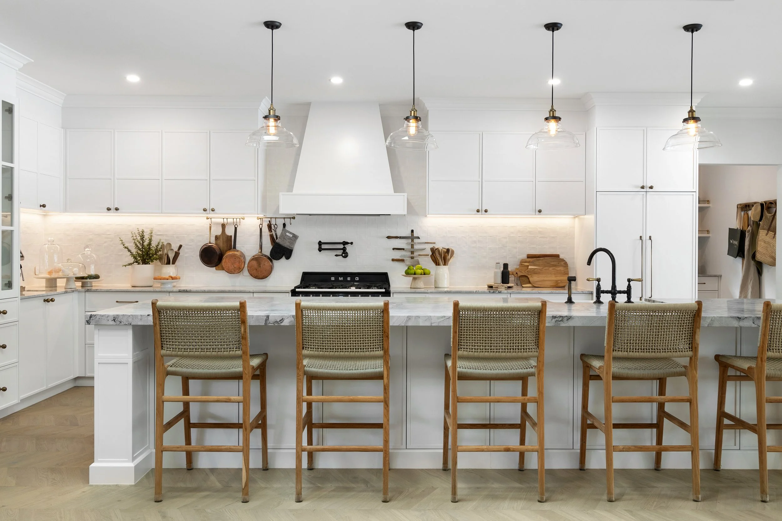 Modern white kitchen with a marble island counter, wooden bar stools, hanging pendant lights, and kitchen utensils hanging on the wall.