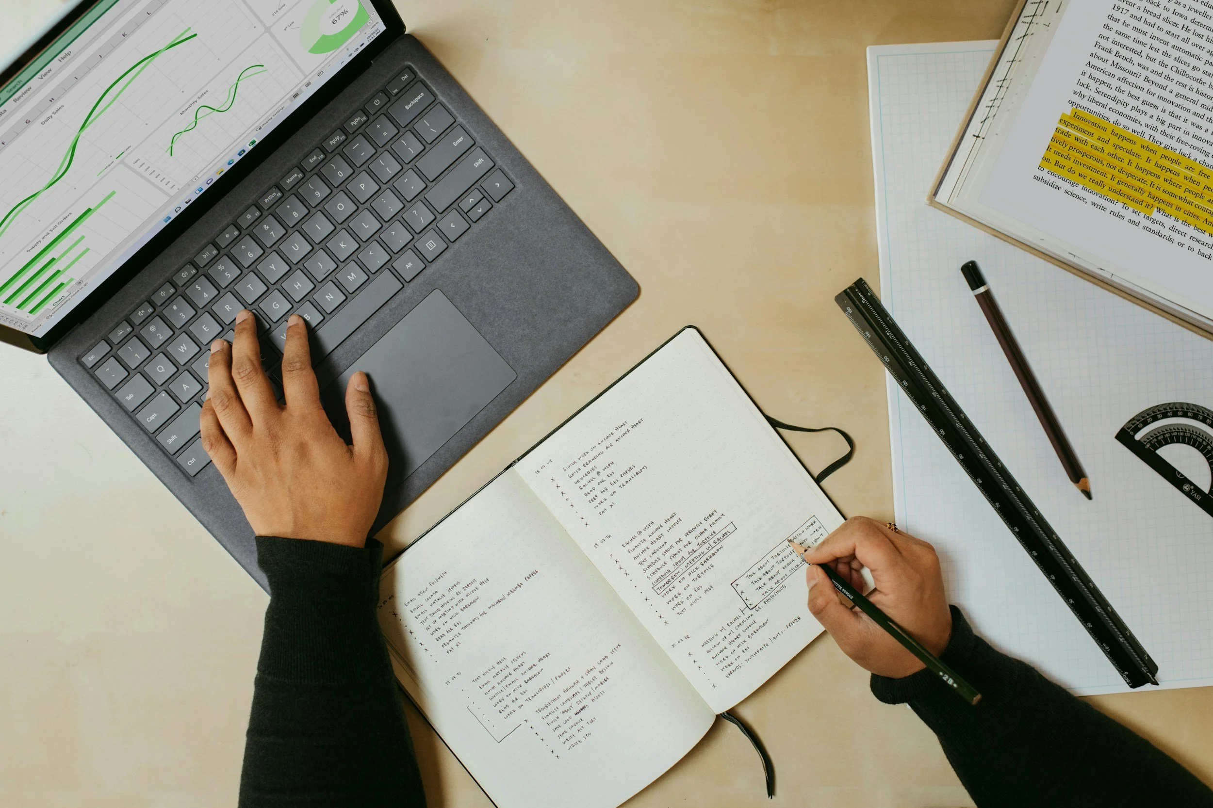 Two people working at a desk with a laptop displaying graphs, and a notebook with handwritten notes, surrounded by rulers, pens, and a book with highlighted text.