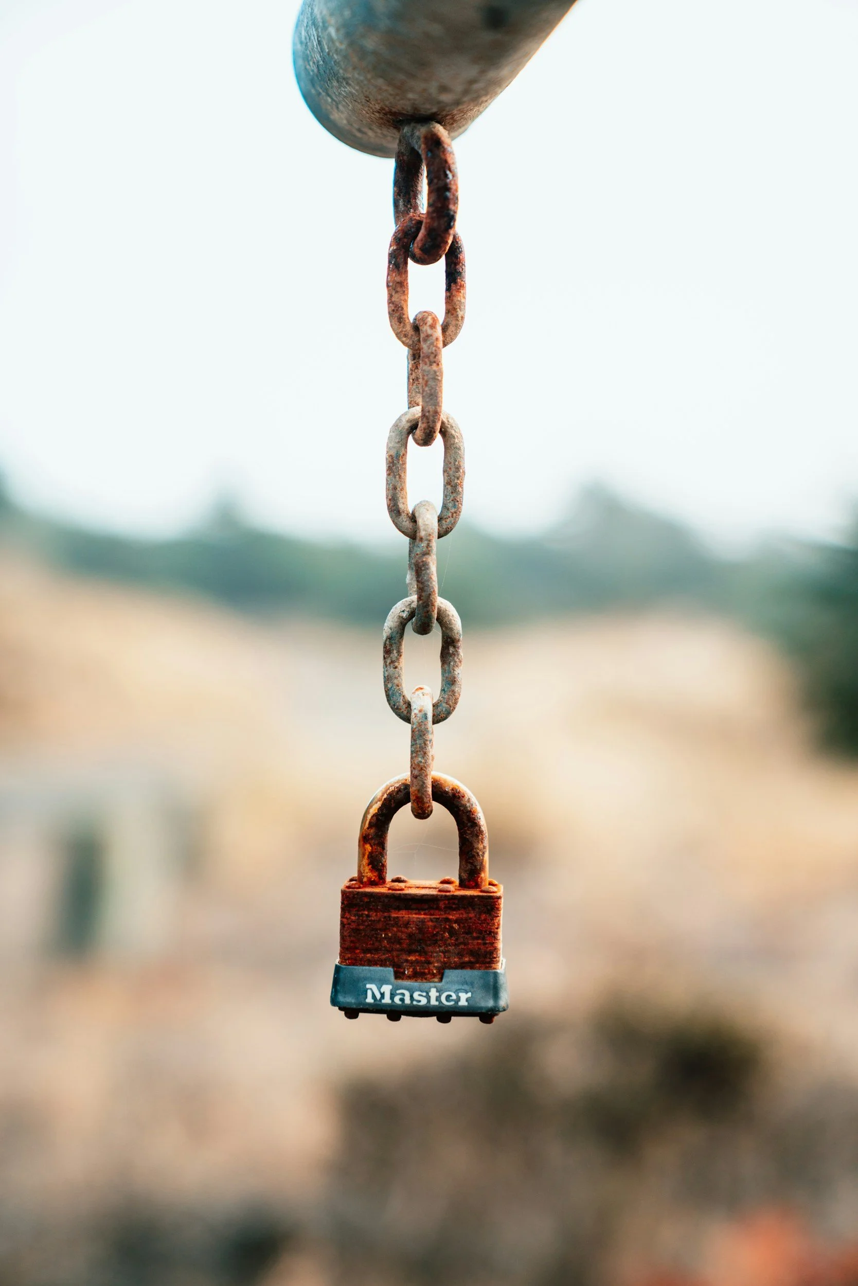 A rusty metal chain with links, hanging from a horizontal surface, with a rusted padlock labeled 'Master' attached at the bottom, against a blurred outdoor background.
