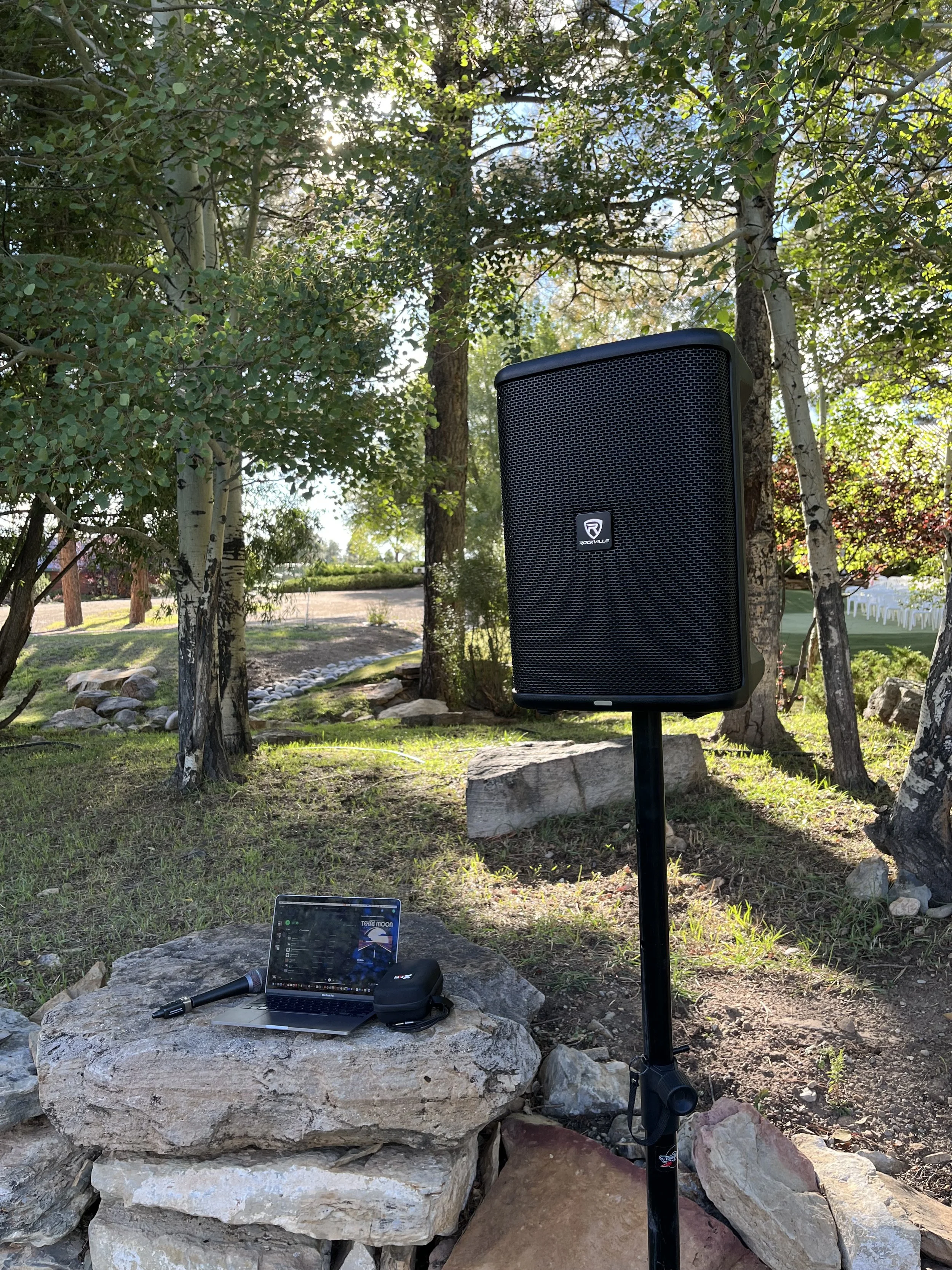 Outdoor scene with a black portable speaker on a stand, a laptop, microphone, and other equipment on rocks, with trees and sunlight in the background.