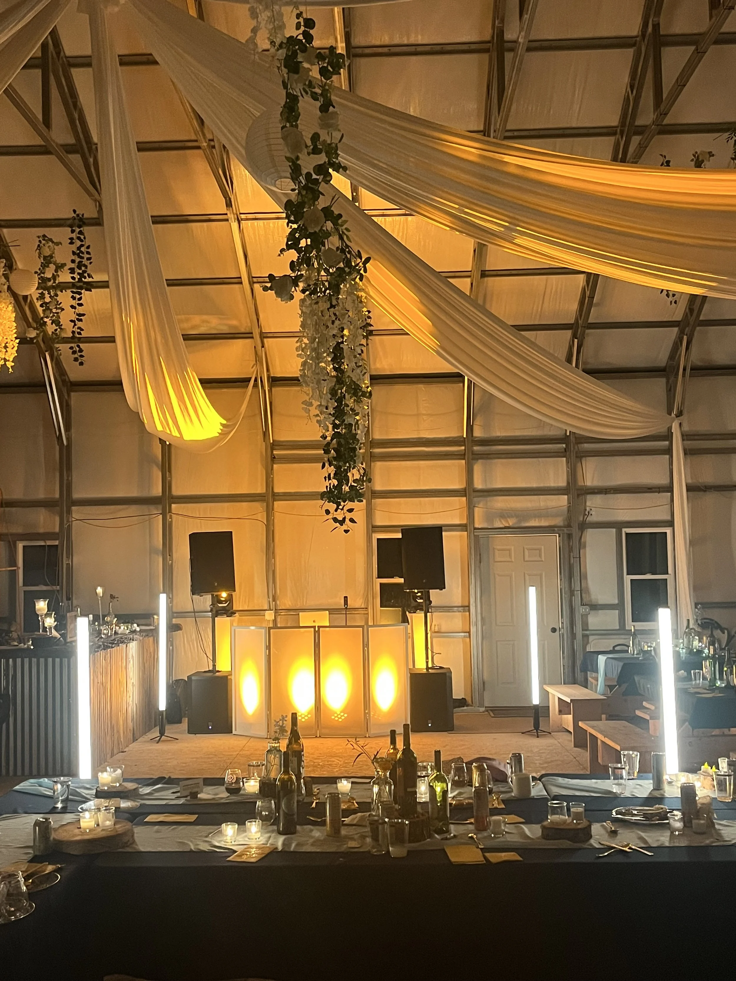 A decorated event space with draped fabric, hanging floral arrangements, warm lighting, and a table set with wine bottles, candles, and glassware for a celebration or ceremony.