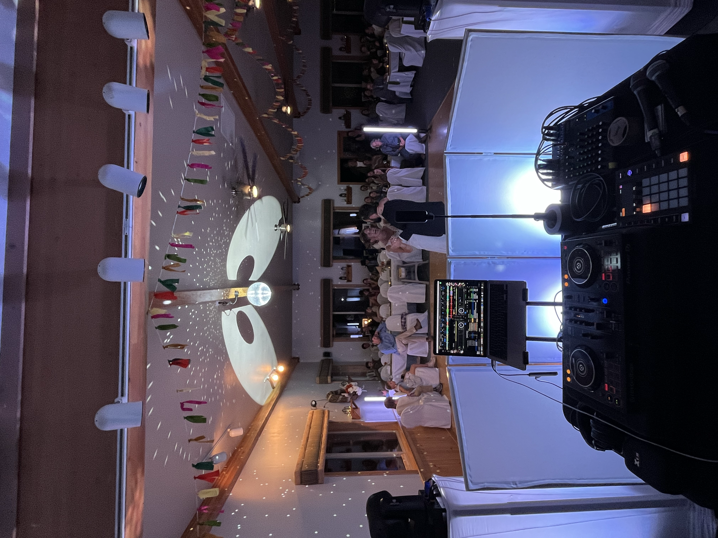 A DJ setup behind a dance floor at a party or event, with a laptop, mixer, and speakers in the foreground. The room is decorated with colorful pennant banners, and there's a disco ball on the ceiling casting light patterns.