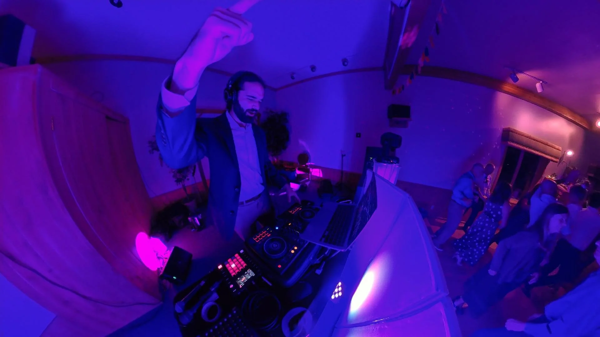 DJ wearing headphones, DJ equipment on a table, people dancing at a party with purple lighting.